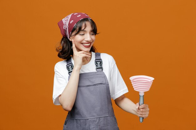 Smiling young female cleaner wearing uniform and bandana holding and looking at plunger while keeping hand on chin isolated on orange background