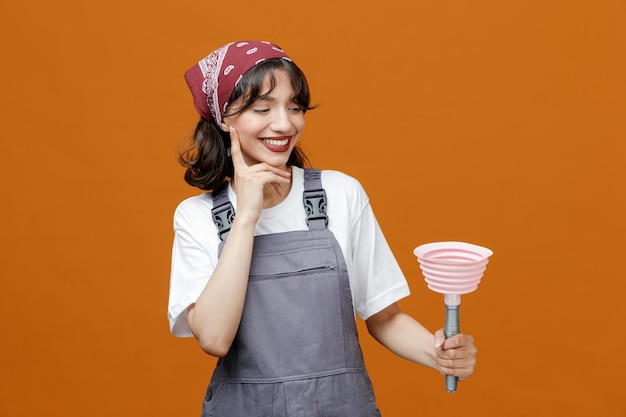 주황색 배경에 격리된 턱에 손을 얹고 플런저를 잡고 유니폼을 입고 반다나를 입고 웃고 있는 젊은 여성 청소부