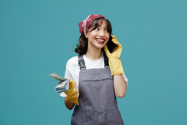 Sorridente giovane donna addetta alle pulizie che indossa bandana uniforme e guanti di gomma che tengono soldi guardando di lato mentre si parla al telefono isolato su sfondo blu