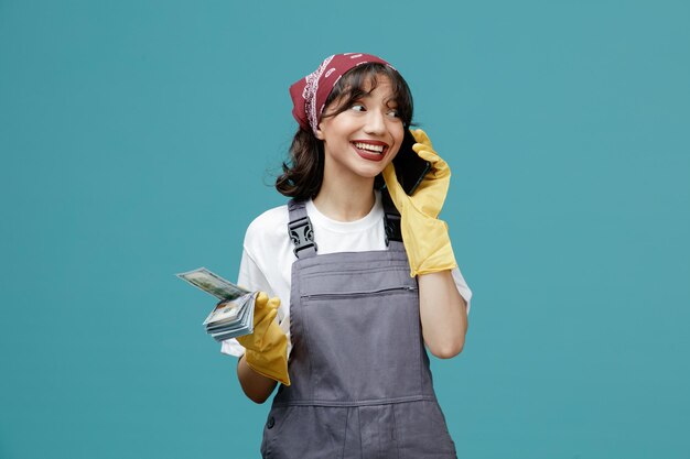 Smiling young female cleaner wearing uniform bandana and rubber gloves holding money looking at side while talking on phone isolated on blue background