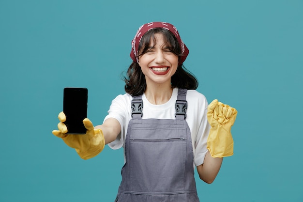 Free photo smiling young female cleaner wearing uniform bandana and rubber gloves stretching mobile phone out towards camera keeping hand in air isolated on blue background