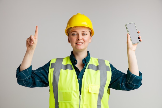 Улыбающаяся молодая женщина-строитель в защитном шлеме и жилете безопасности держит мобильный телефон, указывая вверх