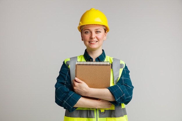 Улыбающаяся молодая женщина-строитель в защитном шлеме и защитном жилете, обнимая блокнот