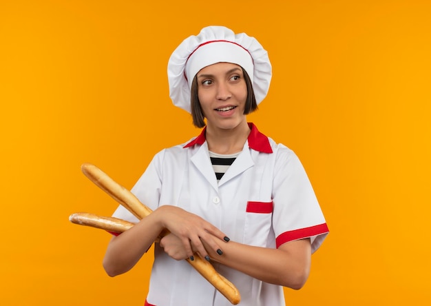 Smiling young female cook in chef uniform holding bread sticks looking at side isolated on orange wall