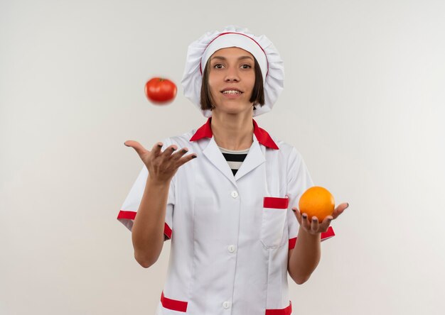 Smiling young female cook in chef uniform holding orange and tossing up tomato isolated on white wall