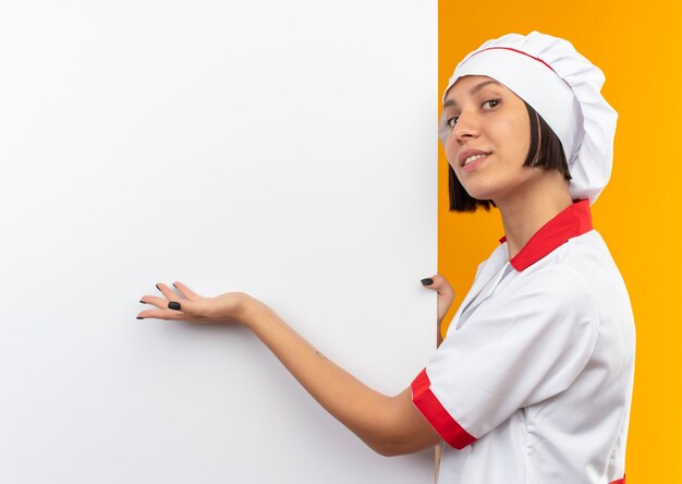 Smiling young female cook in chef uniform standing near white wall holding and pointing with hand at it isolated on orange wall