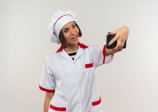 白い壁に分離されたselfieを取るシェフの制服を着た若い女性料理人の笑顔