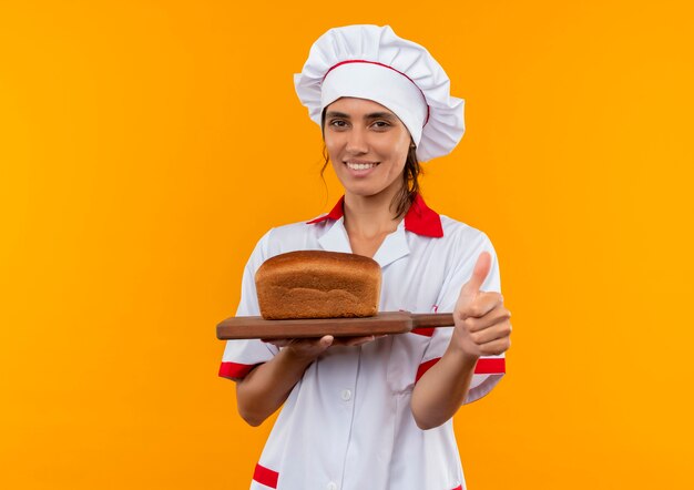 Smiling young female cook wearing chef uniform holding bread on cutting board her thumb up with copy space