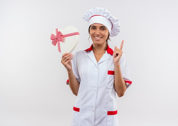 Улыбающаяся молодая женщина-повар в форме шеф-повара держит коробку в форме сердца и указывает вверх с копией пространства