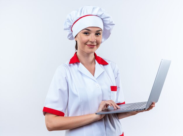 白い壁に分離されたラップトップを保持しているシェフの制服を着て笑顔の若い女性料理人