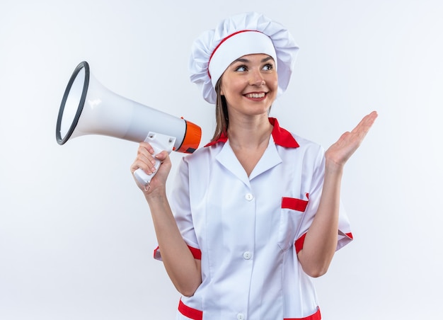 Smiling young female cook wearing chef uniform holding loudspeaker isolated on white background