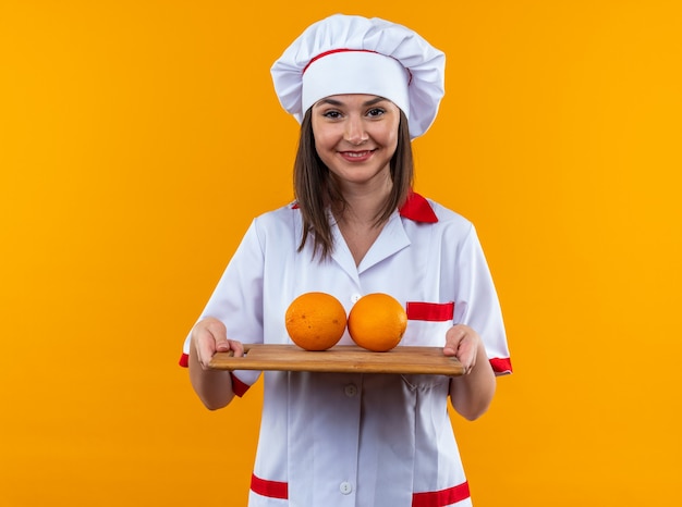 オレンジ色の壁に分離されたまな板にオレンジ色を保持しているシェフの制服を着て笑顔の若い女性料理人