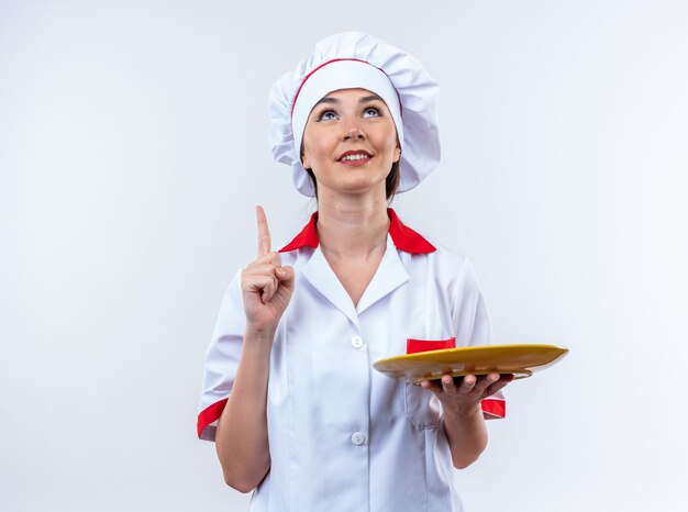 Sorridente giovane cuoca che indossa l'uniforme da chef che tiene la piastra rivolta verso l'alto isolato su sfondo bianco