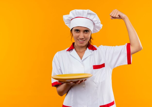 Smiling young female cook wearing chef uniform holding plate and showing strong gesture  with copy space