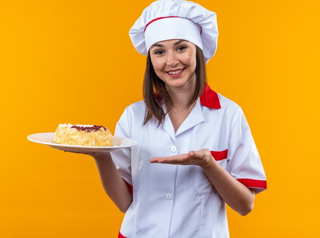 Smiling young female cook wearing chef uniform holding and points at cake on plate isolated on orange background