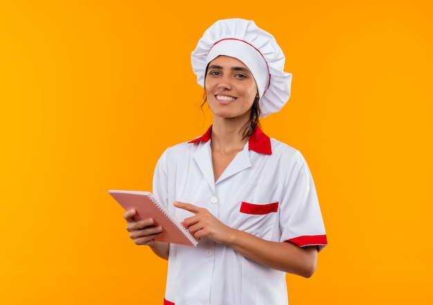 Smiling young female cook wearing chef uniform holding and points notebook  with copy space