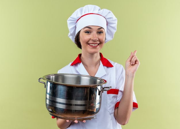 Smiling young female cook wearing chef uniform holding saucepan points at side isolated on olive green background with copy space