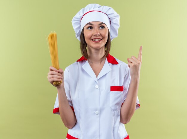 オリーブグリーンの壁に隔離された上でスパゲッティポイントを保持しているシェフの制服を着て笑顔の若い女性料理人