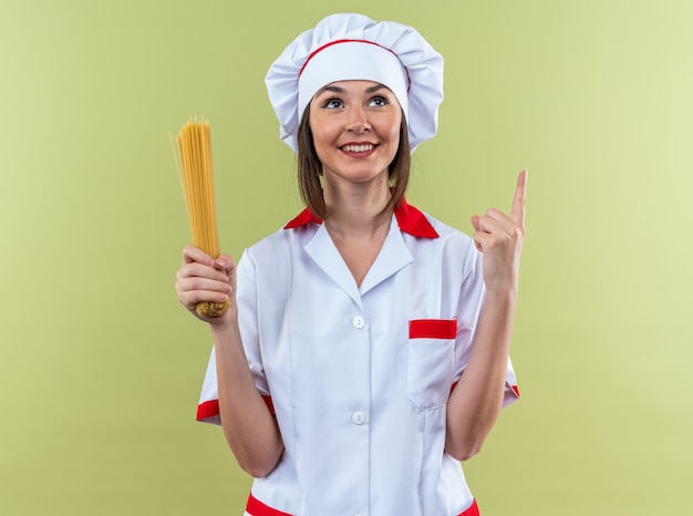 smiling young female cook wearing chef uniform holding spaghetti points at up isolated on olive green wall