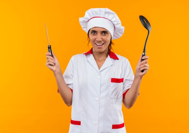 コピースペースでヘラとナイフを保持しているシェフの制服を着て笑顔の若い女性料理人