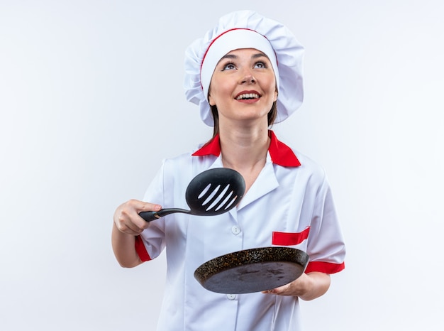 흰 벽에 프라이팬이 달린 주걱을 들고 요리사 유니폼을 입은 웃고 있는 젊은 여성 요리사