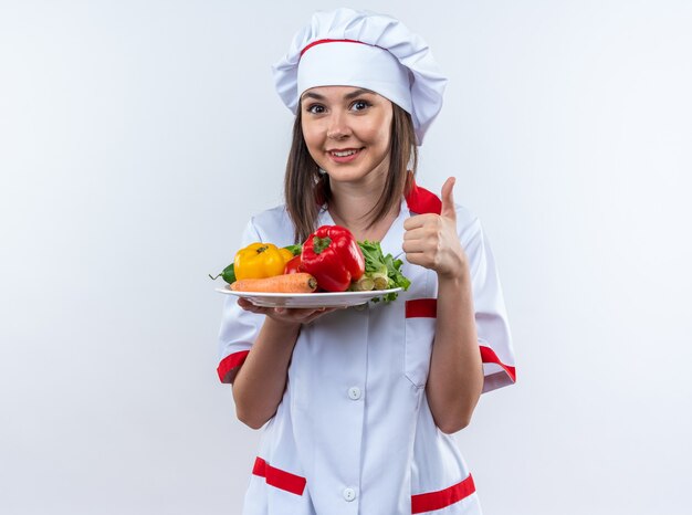 smiling young female cook wearing chef uniform holding vegetables on plate showing thumb up isolated on white wall