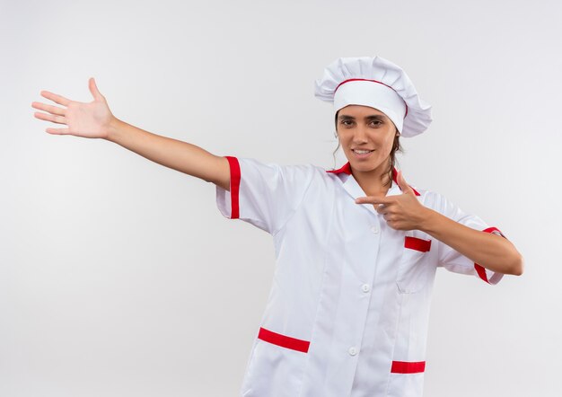 Smiling young female cook wearing chef uniform points to side with hand and finger