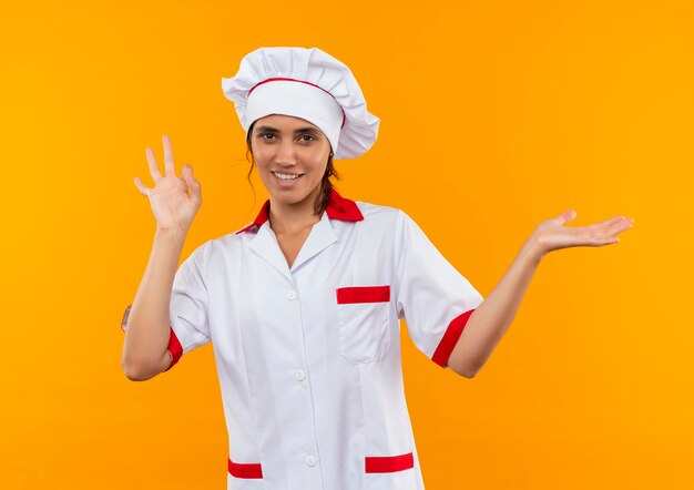 Smiling young female cook wearing chef uniform showing okey gesture and points with hand to side  with copy space