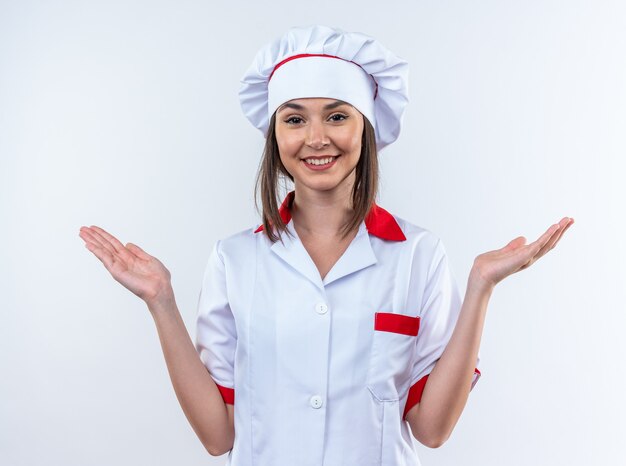 smiling young female cook wearing chef uniform spreading hands isolated on white wall