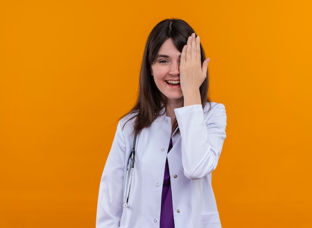 청진 기 의료 가운에 웃는 젊은 여성 의사는 복사 공간이 격리 된 오렌지 배경에 손으로 그녀의 눈을 닫습니다