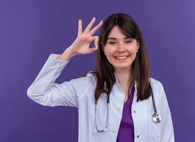 격리 된 보라색 배경에 청진 제스처 확인 의료 가운에 웃는 젊은 여성 의사