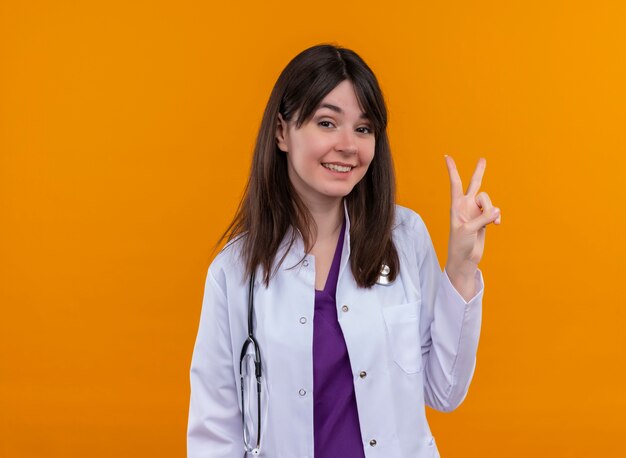 Smiling young female doctor in medical robe with stethoscope gestures victory sign on isolated orange background with copy space