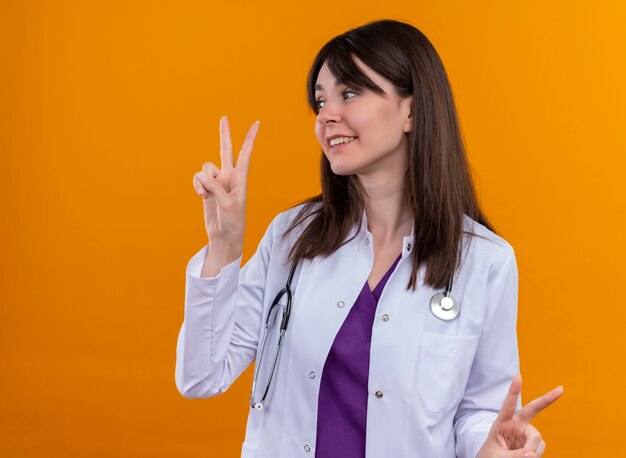 청진 기 제스처와 의료 가운에 젊은 여성 의사 미소 복사 공간이 격리 된 오렌지 배경에 양손으로 승리 기호