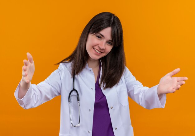 Smiling young female doctor in medical robe with stethoscope holds both hands open on isolated orange background with copy space