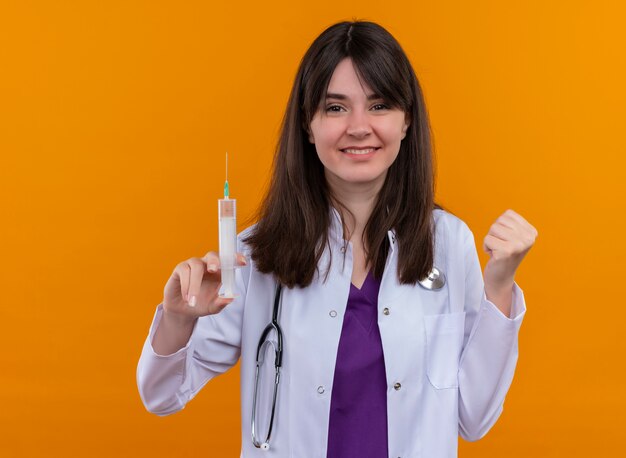 Smiling young female doctor in medical robe with stethoscope holds syringe and keeps fist up on isolated orange background with copy space