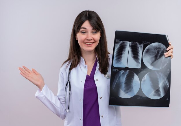 Smiling young female doctor in medical robe with stethoscope keeps empty hand up and holds x-ray shot on isolated white background with copy space