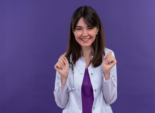 Smiling young female doctor in medical robe with stethoscope keeps hands closed on isolated violet background with copy space