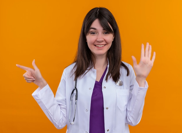 Sorridente giovane dottoressa in abito medico con lo stetoscopio punta a lato e tiene la mano su sfondo arancione isolato