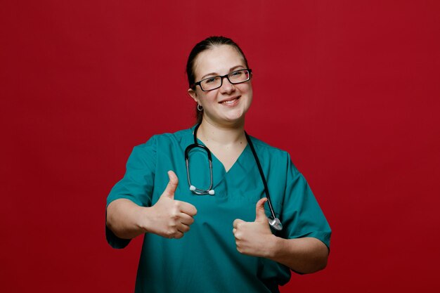 Smiling young female doctor wearing glasses uniform and stethoscope around her neck looking at camera showing thumbs up isolated on red background