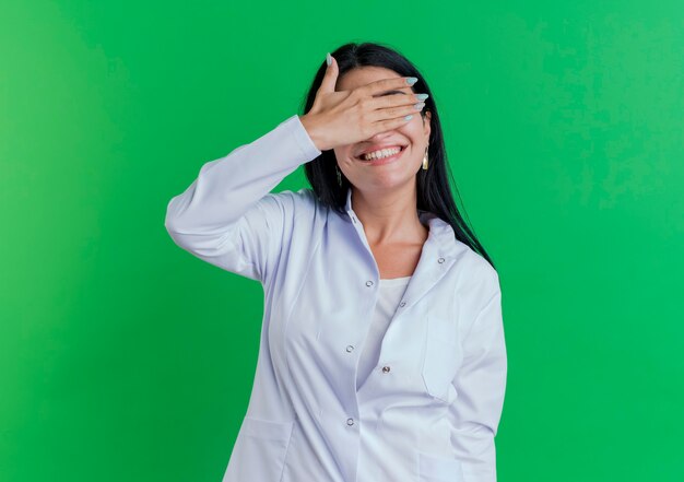Smiling young female doctor wearing medical robe covering eyes with hand isolated on green wall with copy space