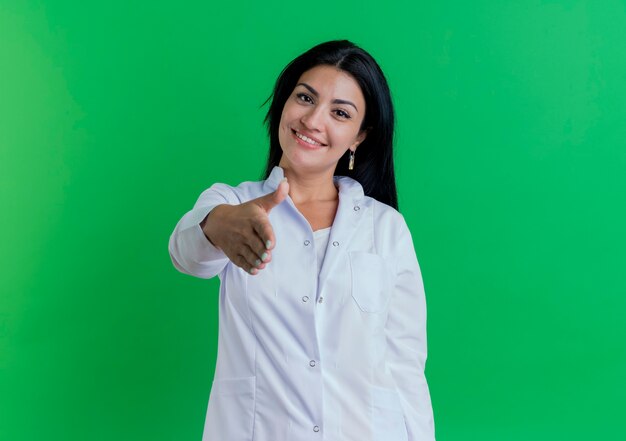 Smiling young female doctor wearing medical robe doing hi gesture  greeting someone isolated on green wall with copy space