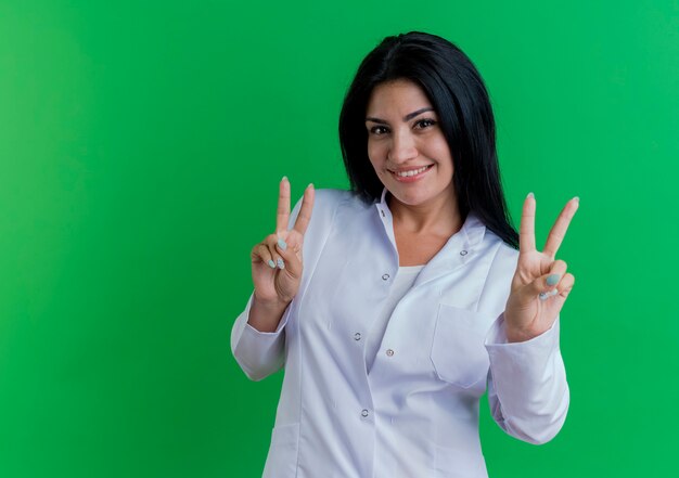 Smiling young female doctor wearing medical robe  doing peace sign isolated on green wall with copy space