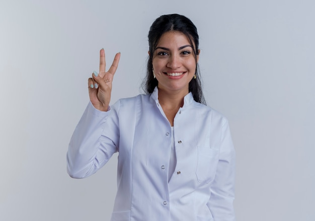 Smiling young female doctor wearing medical robe doing peace sign  isolated on white wall with copy space