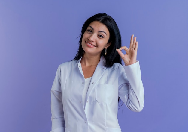 Smiling young female doctor wearing medical robe looking doing ok sign 