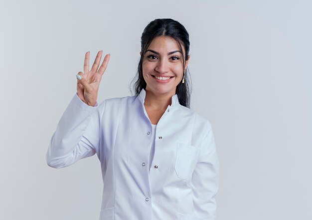 Smiling young female doctor wearing medical robe looking and showing three with hand isolated