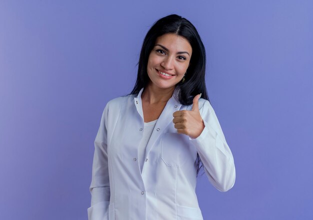 Smiling young female doctor wearing medical robe looking showing thumb up 