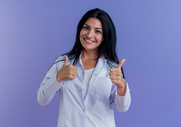 Smiling young female doctor wearing medical robe looking showing thumbs up 