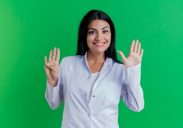 Улыбающаяся молодая женщина-врач в медицинском халате, показывающая девять с руками, изолированными на зеленой стене с копией пространства