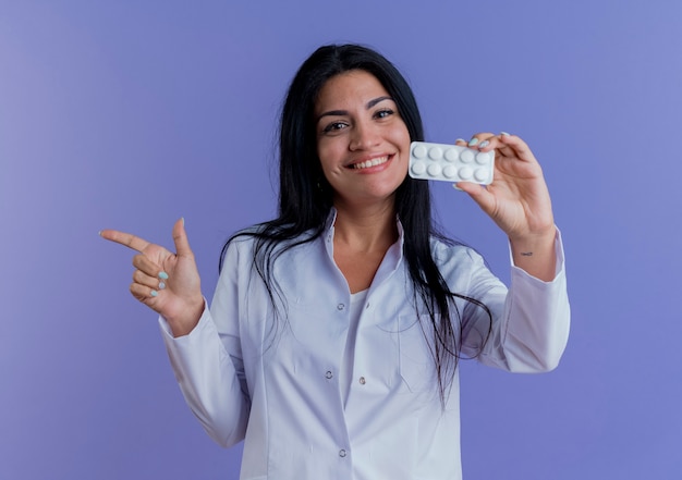 Smiling young female doctor wearing medical robe showing pack of medical tablets, looking pointing at side 