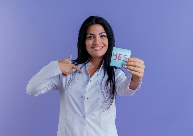 Smiling young female doctor wearing medical robe showing yes note, looking pointing at herself 